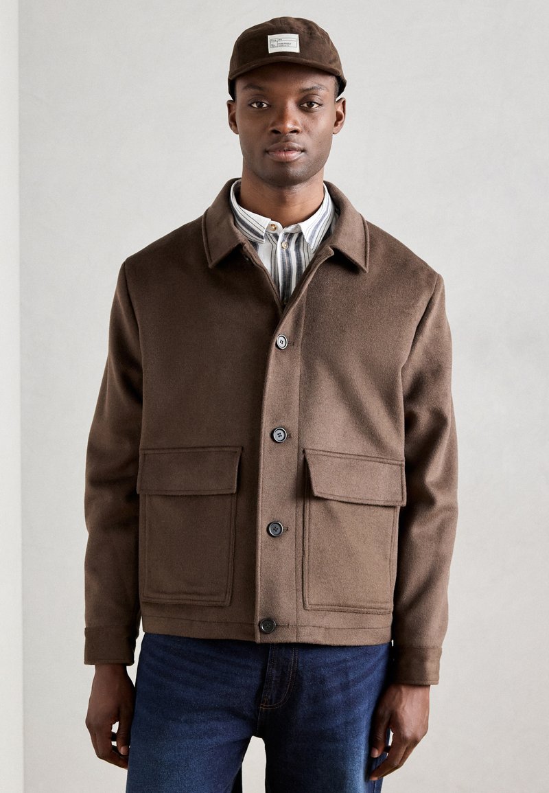 Giacca in misto lana marrone con colletto, bottoni frontali e due grandi tasche a toppa. Abbinata a una camicia a righe leggera e a dei jeans.
