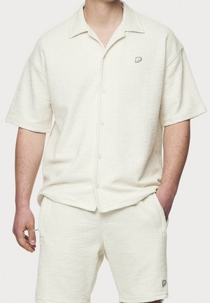 Hombre vestido con una camisa corta de botones de manga corta en textura crema a juego con pantalones cortos, ambos con pequeños logotipos de "P" en el pecho y en la pierna izquierda.