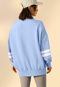 Felpa oversize blu chiaro con strisce bianche sulle maniche. Tessuto morbido, polsini e orlo a coste. Abbinata a leggings grigi con texture.