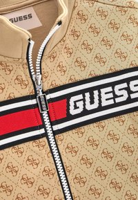 Veste zippée beige avec un motif en losanges et un logo subtil, rayures noires et blanches avec un accent rouge, et fermeture éclair argentée.