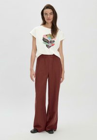 Witte katoenen t-shirt met een kleurrijk grafisch ontwerp op de voorkant, gecombineerd met losse, donkerrode, wijde broek en zwarte schoenen.