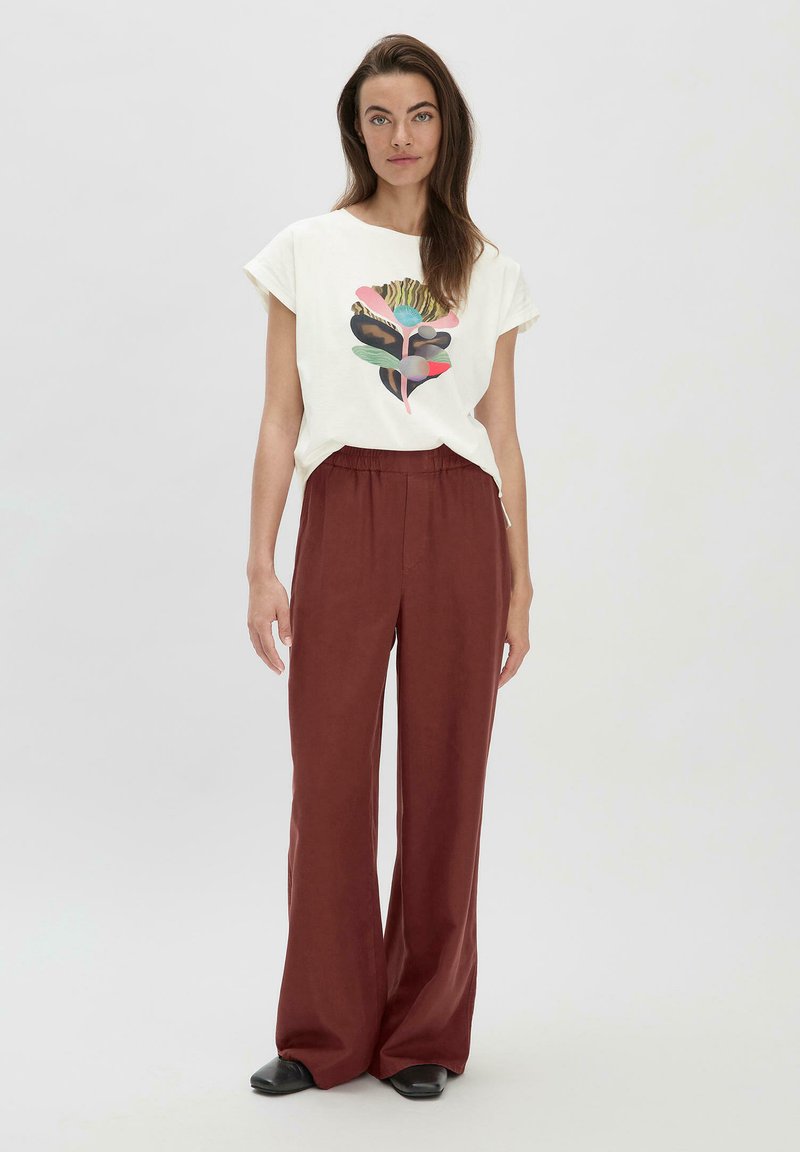 Witte katoenen t-shirt met een kleurrijk grafisch ontwerp op de voorkant, gecombineerd met losse, donkerrode, wijde broek en zwarte schoenen.