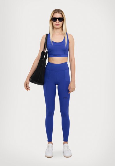 Modrý sportovní set skládající se z croptopu a legín s vysokým pasem. Materiál je hladký s těsným střihem; zahrnuje černou tašku.