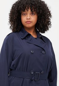 Marinblå trenchcoat gjord av slät tyg, med dubbelknäppt knappning, bälte och en klassisk krage.