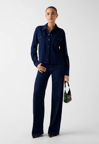 Veste en denim bleu marine avec deux poches poitrine, associée à un pantalon large assorti. Le modèle tient un petit sac à main vert. Texture lisse.