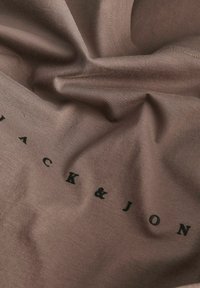 Gros plan sur un tissu brun froissé avec un texte imprimé en noir « JACK & JON » partiellement visible à la surface.