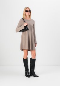 Robe beige à manches longues en tissu doux, associée à des bottes noires hautes jusqu'aux genoux et à un petit sac à main noir. Le modèle porte des lunettes de soleil sombres.