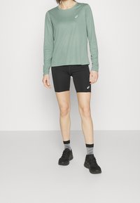 Langärmliges, hellgrünes Sportshirt kombiniert mit schwarzen Fahrradhosen, versehen mit einem Logodetail. Das Model trägt schwarze Schuhe und graue, gemusterte Socken.