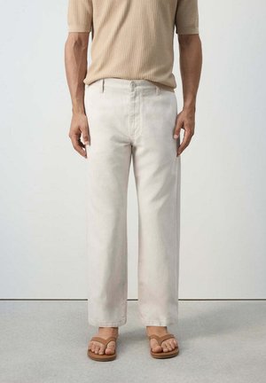 Persona que lleva pantalones beige claro de pierna ancha, camiseta de manga corta de punto waffle color marrón claro y sandalias marrones de cuero con tira entre los dedos, de pie sobre suelo gris.