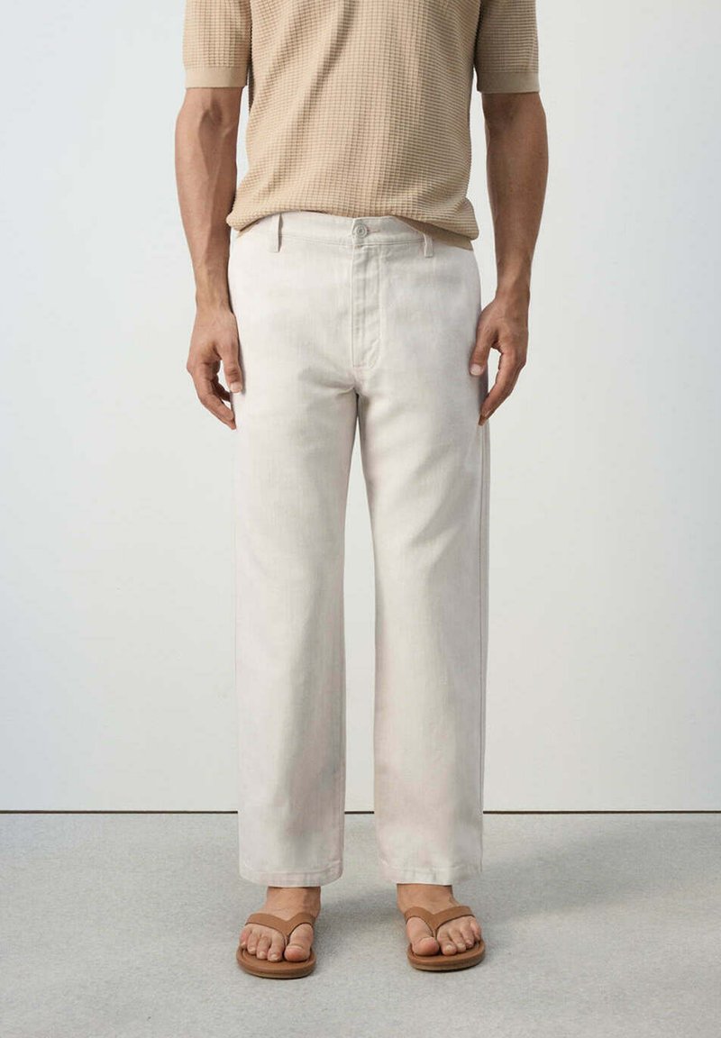 Personne portant un pantalon ample beige clair, un t-shirt à manches courtes en maille gaufrée couleur tan, et des sandales en cuir marron à lanière entre les orteils, debout sur un sol gris.
