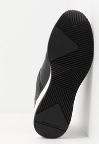 Semelle d'une basket Michael Kors noir et blanc avec des motifs de grip texturés et le nom de la marque au talon sur fond blanc.