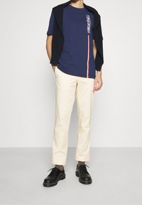 Námořnické tričko s krátkým rukávem a bílou grafickou motivací, doplněné krémovými kalhotami. Černé boty dokončují tento outfit. Strukturovaný svetr přehozený přes ramena.