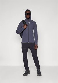 Tamno siva uska zip-up majica s kapuljačom, uparena s crnim hlačama i taktičnim crnim čizmama. Model nosi crni ruksak.