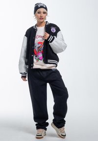 Black and white varsity jacket, colorful graphic sweatshirt, dark loose-fit jeans, beige sneakers with black accents, and a black beanie.