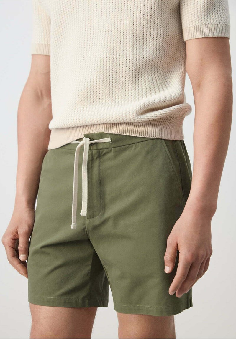 Homme portant un short vert à cordon de serrage et un pull fin en maille texturée crème à manches courtes, les mains détendues le long du corps.
