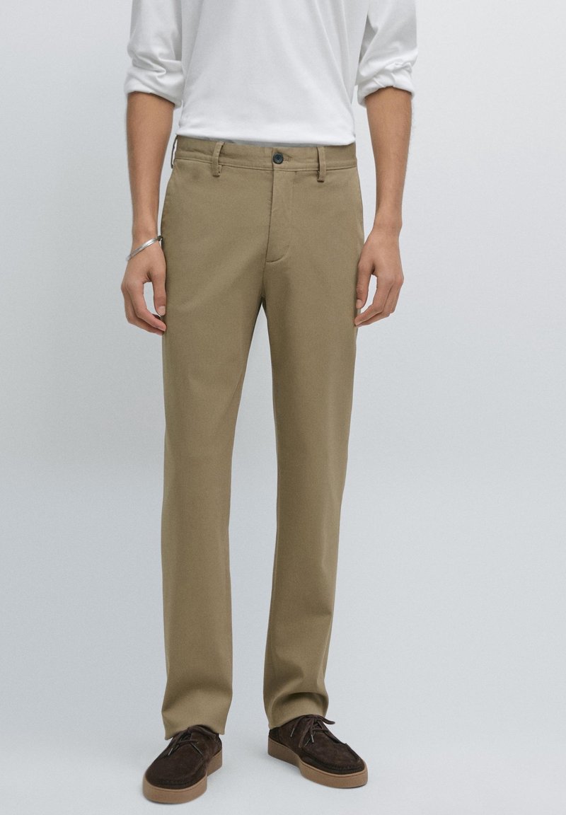 Homme portant un pantalon beige à jambes droites, une chemise blanche à manches longues, un bracelet argenté et des chaussures à lacets marron foncé, sur un fond uni.