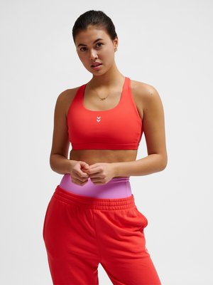 Junge Frau trägt roten Sport-BH und rote Jogginghose über pinken Leggings, steht vor weißem Hintergrund.