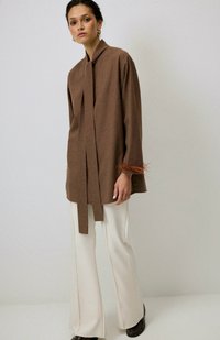 Camicia a maniche lunghe marrone con colletto e nodo frontale, con dettagli testurizzati ai polsini; abbinata a pantaloni a palazzo color crema.