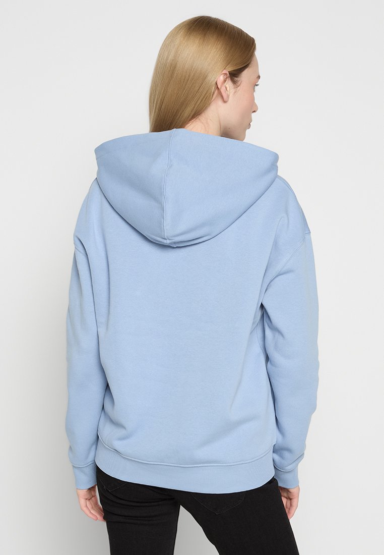 Jasnoniebieska bluza z kapturem, wykonana z miękkiego materiału, z dużym kapturem, luźnym krojem oraz ściągaczami na rękawach i dolnym końcu. Widok z tyłu ukazuje minimalną ilość szwów.