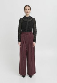 Camicia nera in pizzo a maniche lunghe con apertura frontale a bottoni e collo alto, abbinata a pantaloni larghi a righe bordeaux e scarpe nere a punta.