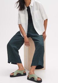Chemise blanche légère à boutons avec deux poches avant, associée à un pantalon ample vert foncé et des sandales vertes à lanières croisées sur un piédestal en bois.