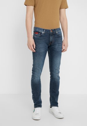 Man wearing slim-fit blue jeans, white sneakers, and a brown short-sleeve shirt, standing against a plain light background.