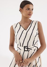 Sukienka w kształcie litery V w kremowym materiale z czarnymi pionowymi paskami, z belką w talii i guzikami. Teksturowana powierzchnia o gładkim wykończeniu.