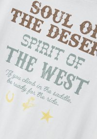 Valge puuvillane t-särk, millel on kulunud mitmevärviline tekst: pruun, roheline ja kollane, koos lääne temaatiliste ikoonidega, nagu hobuseraud, täht ja kauboi.