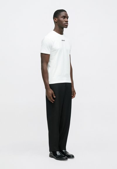 Biała koszulka z krótkim rękawem z logo "VERSACE", zestawiona z czarnymi spodniami o szerokich nogawkach i czarnymi butami, na jednolitym tle.