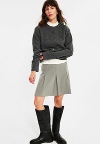Frau in einer dunkelgrauen, geknöpften Strickjacke, weißem Rollkragenpullover, grauem, gefaltetem Rock und schwarzen knielangen Stiefeln, stehend vor einem weißen Hintergrund.