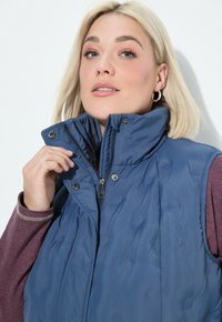 Blonde Frau, die den Kragen einer blauen, gepolsterten Weste anpasst, die über einem langärmeligen kastanienbraunen Hemd getragen wird, und leicht nach oben blickt.