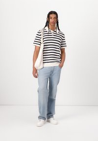 Stribet strikket polo-shirt i marineblå og creme, afslappede slidte blå jeans og hvide sneakers. Bærer en kanvas tote bag.