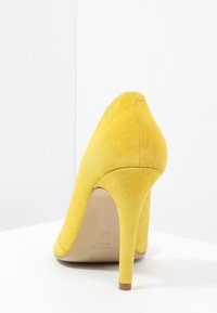 Chaussure à talons hauts en daim jaune avec un bout pointu et un talon fin, présentant une texture lisse et un design minimaliste.
