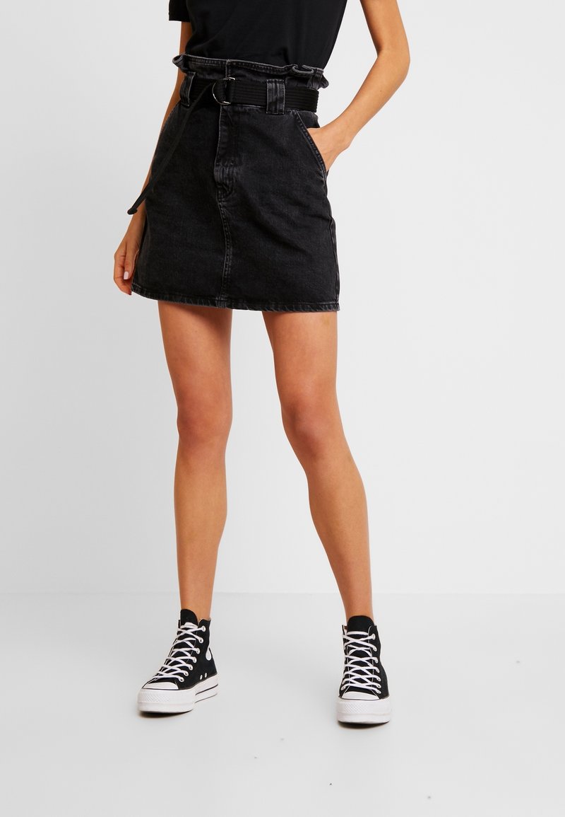 Kvinna som bär en svart hög midja jeanskjol med en volangbälte och svarta hög-top sneakers med vita snören, stående med ena handen i fickan.