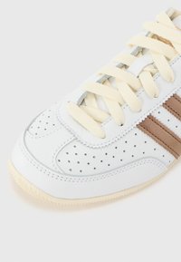 Adidași din piele albă, cu detalii perforate, accente din suédă bej, șireturi de culoare bej deschis și o talpă din cauciuc texturată.