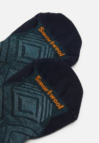 Socken mit einem dunkelblauen Grundton, verziert mit einem geometrischen tealfarbenen Muster und orangefarbenem "Smartwool"-Branding am oberen Rand. Hergestellt aus weichem Material.