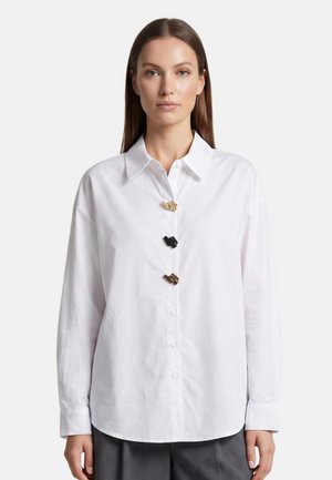 Femme portant une chemise blanche à manches longues avec trois boutons décoratifs uniques en or et noir, associée à un pantalon gris foncé, de face.