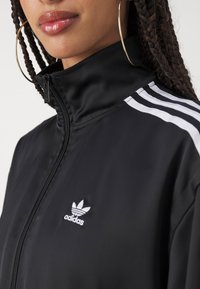 Chaqueta negra con cremallera hecha de una tela suave, con acentos de tres rayas blancas de Adidas en las mangas y un pequeño logo blanco en el pecho.