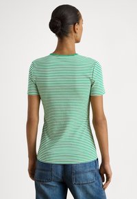 Žena s vlasmi zviazanými do drdolu, oblečená v zelenej a bielo pruhovanej krátkosrievkovej košeli a modrých rifliach, stojaca chrbtom k jednoliatemu pozadiu.