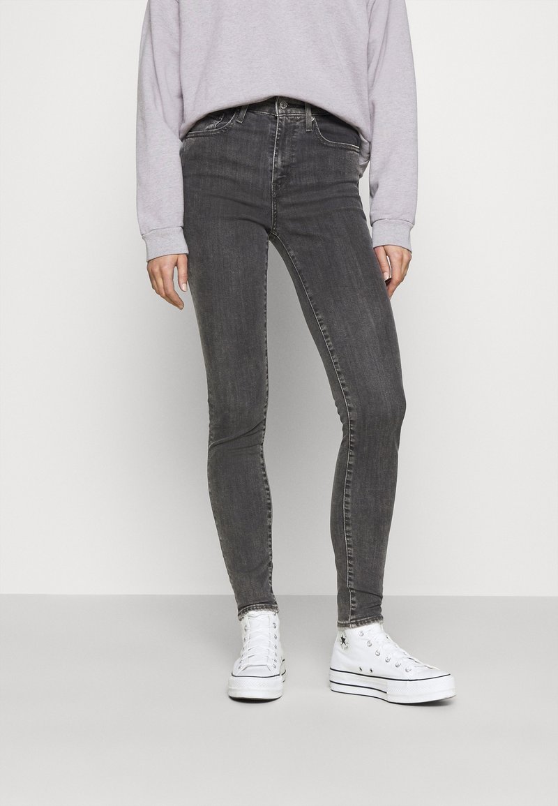 Personne portant un jean skinny gris foncé, des baskets blanches montantes et un haut à manches longues gris clair, debout devant un fond uni.