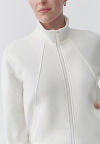 Giacca bianca con zip e colletto alto, realizzata in tessuto morbido. Presenta dettagli delle cuciture e tasche laterali. Design classico e minimalista.