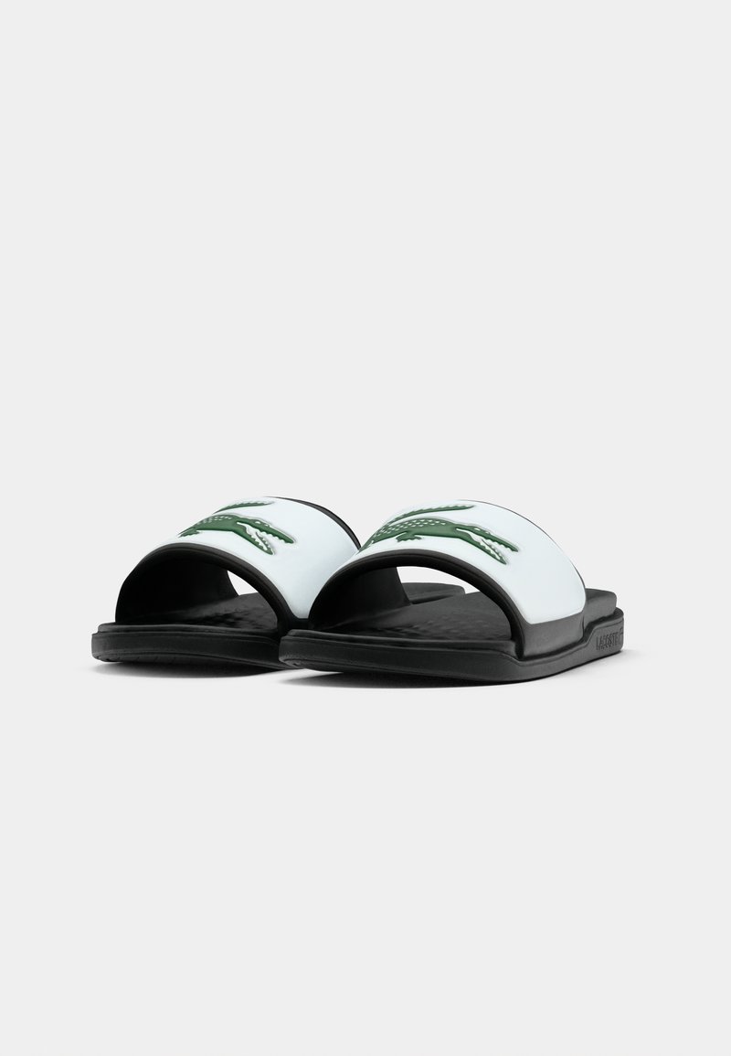 Des sandales à glissière noires et blanches avec un logo de crocodile vert sur la sangle blanche, montrées sous un angle avant bas sur fond blanc.