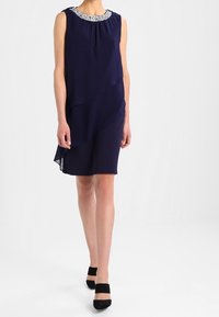 Robe sans manches bleu marine avec un design en couches, dotée d'un col à sequins. Portée avec des talons noirs à bride de cheville. Tissu lisse, silhouette élégante.