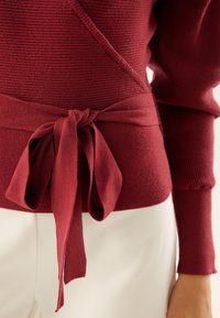 Bordeauxfarbener Wickelpullover mit geripptem Struktur, ausgestattet mit einem gebundenen Taillendetail und langen Ärmeln, kombiniert mit cremefarbenen Hosen.