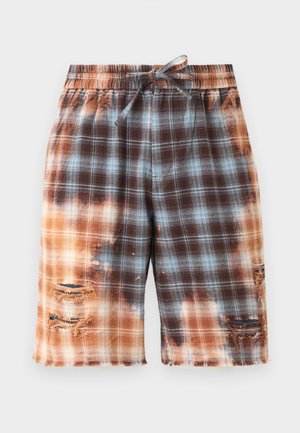 Homme portant des lunettes de soleil noires, une chemise blanche boutonnée, un short à carreaux tie-dye avec des détails usés, des chaussettes blanches et des bottes rouge foncé.