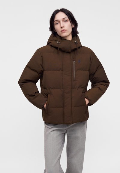Personne portant une doudoune marron avec une capuche et un pantalon gris clair, debout les mains dans les poches devant un fond uni.