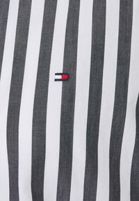 Stripet stof med skiftende sorte og hvide lodrette linjer. Et lille broderet logo i rød og blå er til stede.