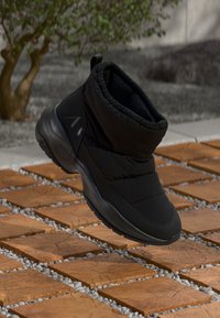 Schwarzer, bauschiger Knöchelstiefel mit einem strukturierten Obermaterial, gepolstertem Kragen und einer dicken Gummisohle. Mit einem erhabenen Logo an der Seite.