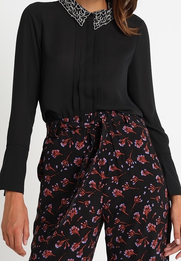 Femme portant un blouse noire avec des broderies florales blanches sur le col et un pantalon noir avec des motifs floraux rouges et violets.