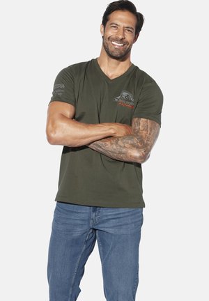 Olivgrünes V-Ausschnitt-T-Shirt mit kurzen Ärmeln, das ein bedrucktes Design auf der linken Schulter und der Brust zeigt. Trägt dazu eine hellblaue Jeans.